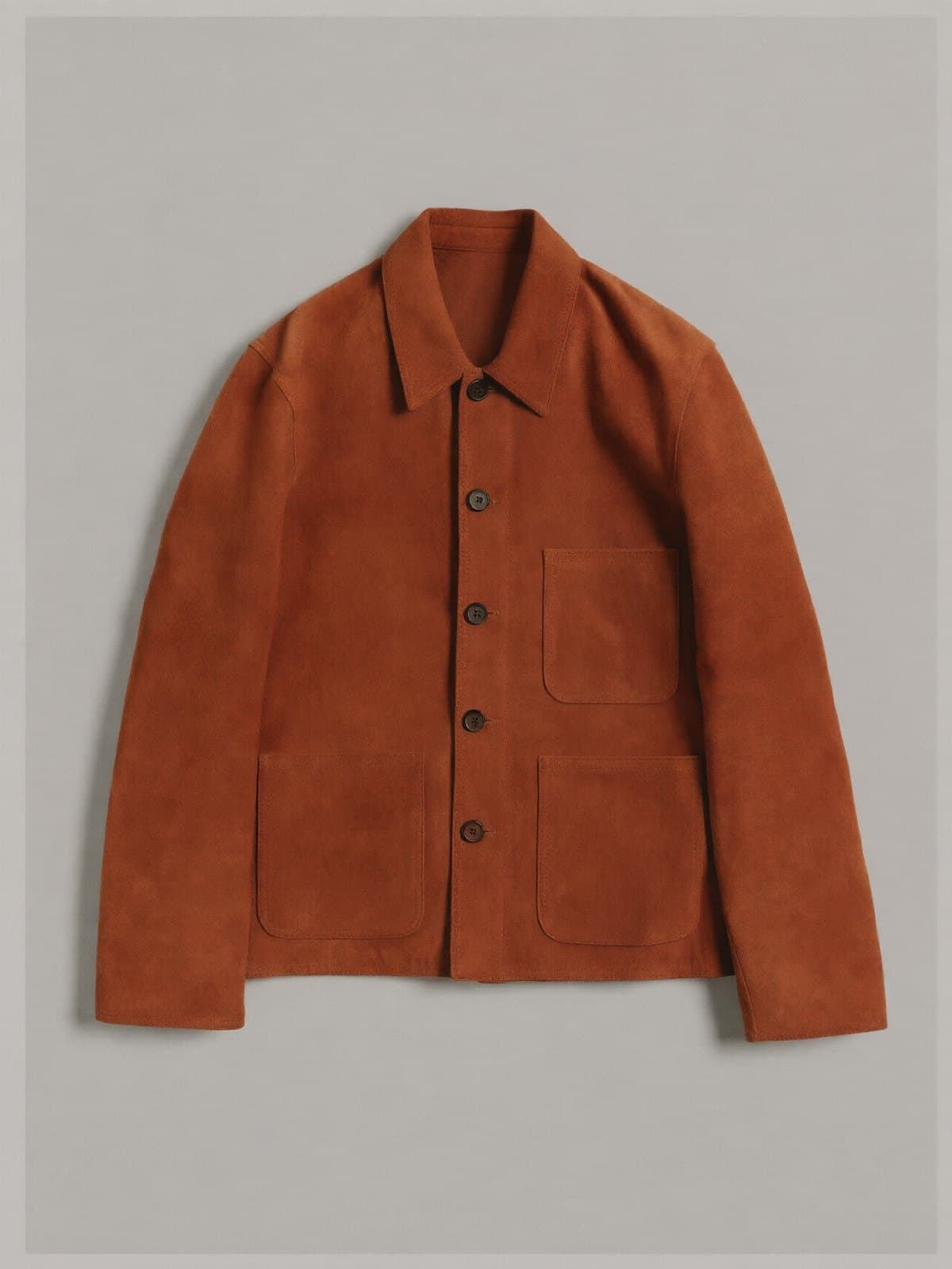 A rust suede chore jacket with patch pockets laid flat on light grey paper.